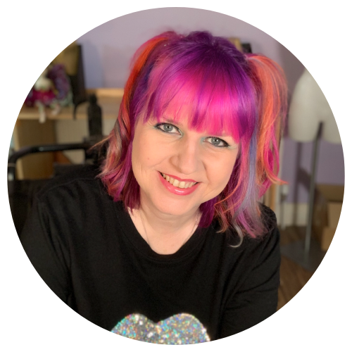 Colour photo of Nicky in her wheelchair in her sewing room. Nicky has pink, purple and orange hair and is smiling at the camera.