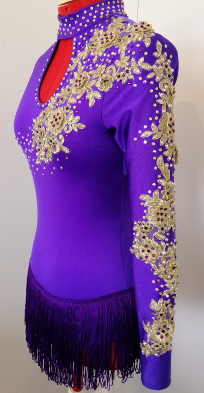 TWA 1 - Tutus with Aroha Photograph of a purple dance costume by Tutus With Aroha featuring a purple fringe skirt and gold lace appliqués and crystals. The appliqués are across the left shoulder and down the full length of the left arm. The crystals surround the appliqués and continue across the body and neckline.